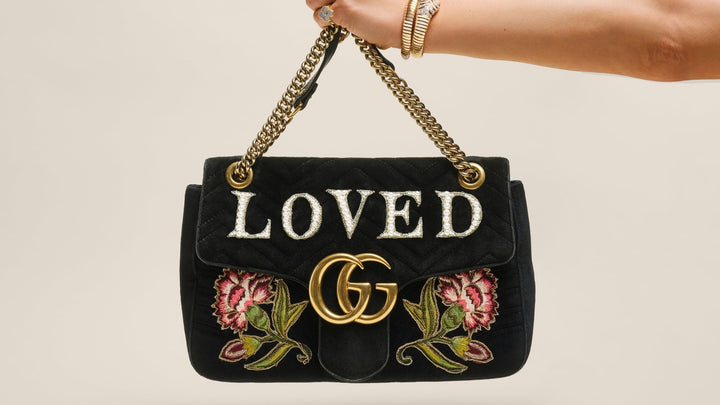 A hand wearing gold jewelry holds up a black velvet Gucci GG Marmont