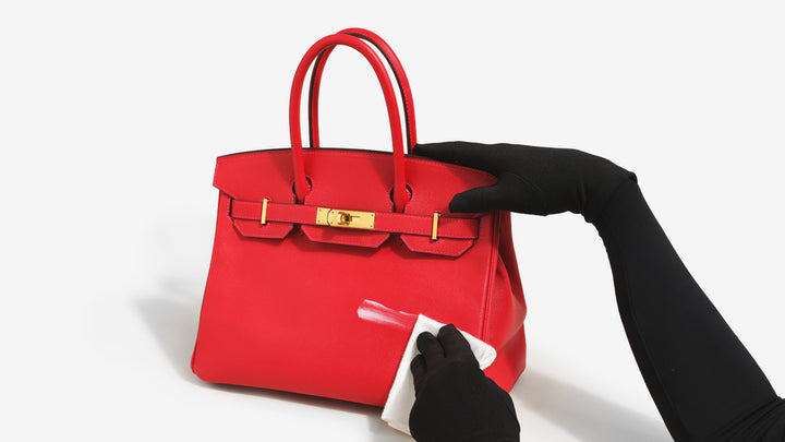 A gloved black hand cleans a red Hermes Birkin using ReNew by FASHIONPHILE