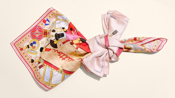 A colorful Louis Vuitton scarf and a light pink Hermes scarf are tied together in a tight knot in the center against a plain light background.