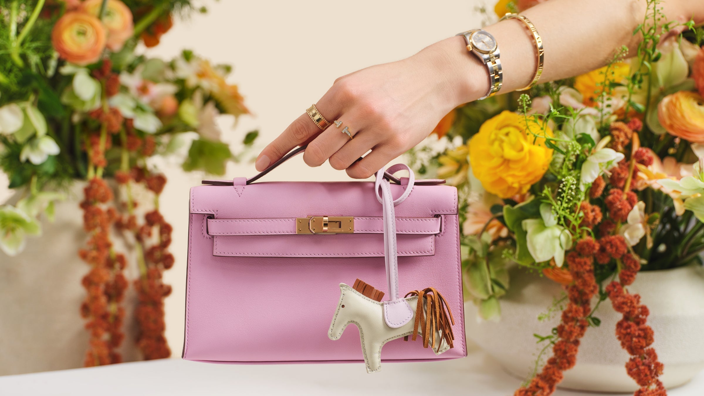 A hand holding a Hermes Swift Kelly Pochette Clutch in Mauve Sylvestre with an Hermes bag charm.