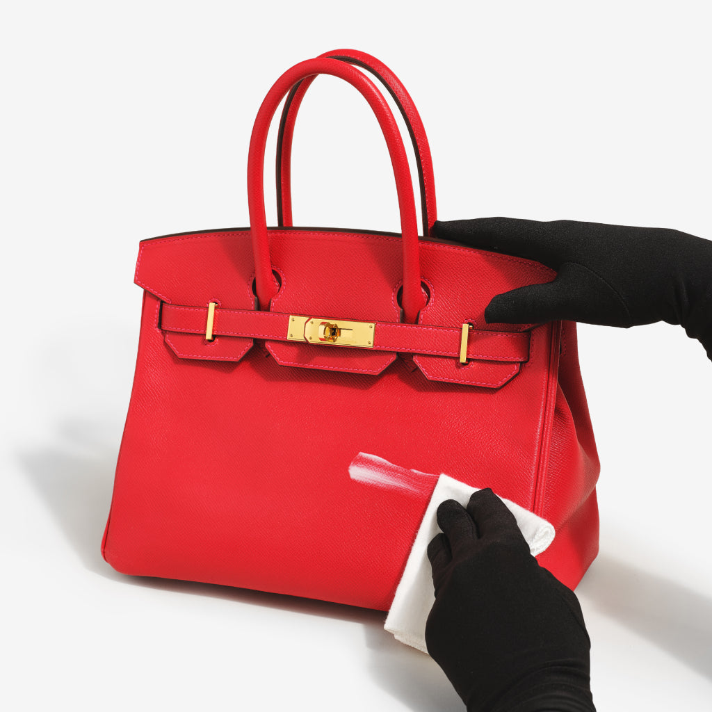 A gloved hand uses a premium microfiber cloth to gently buff handbag cleaner into a Birkin. The cloth and the cleaner are part of the ReNew Handbag Cleaning Kit.
