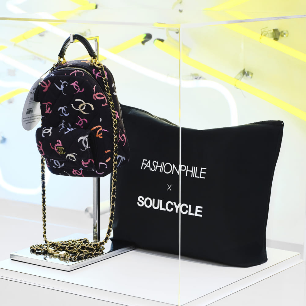 FASHIONPHILE x SoulCycle Sweat Bag in a display case with a Chanel backpack