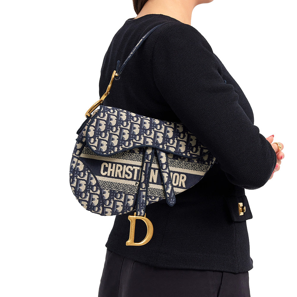 this is an image by fashionphile of a model holding a blue and white oblique christian dior classic saddle bag as a shoulder bag