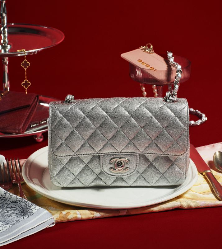 one Chanel Metallic Calfskin Quilted Mini Rectangular Flap Silver on a place setting with party glasses and trays as props holding Van Cleef Alhambra bracelet, a pink Gucci Card Holder and a red Saint Laurent card holder