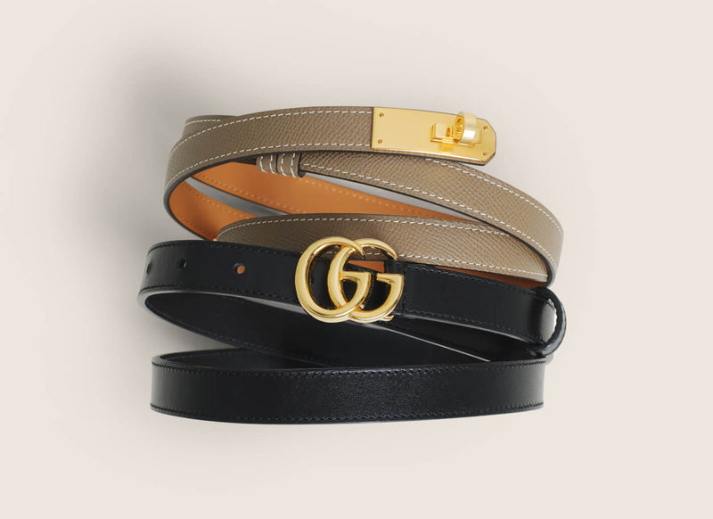 Two leather belts stacked on a light beige background: a black belt with a gold double-G buckle, a taupe belt with a gold buckle, and a tan belt with a gold buckle.