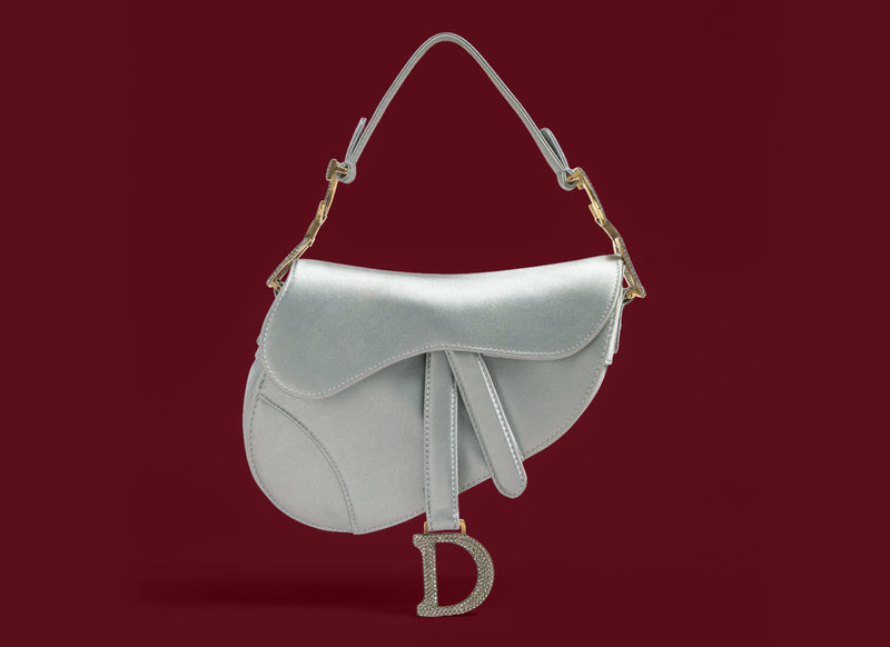 A silver leather saddle bag with gold-tone hardware and a rhinestone-embellished 'D' charm, shown against a solid dark red background.