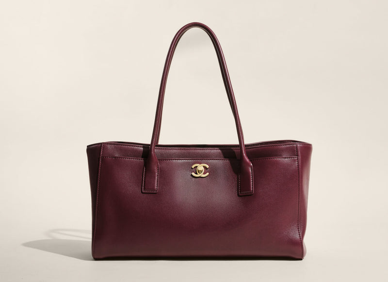 A dark burgundy leather tote bag with double top handles and a gold-tone interlocking CC logo on the front, characteristic of Chanel. The bag is photographed against a clean, solid, warm off-white background.