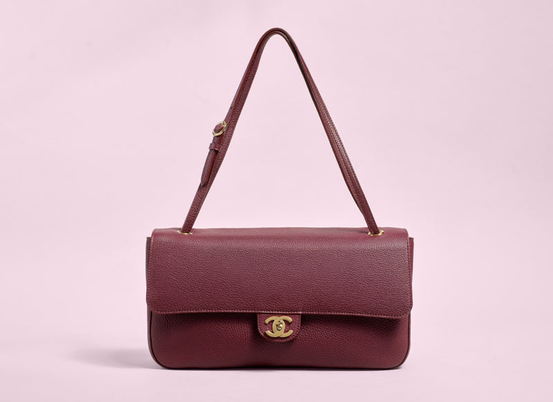 A dark burgundy leather shoulder bag with a gold-tone interlocking CC logo clasp, displayed against a solid light pink background.