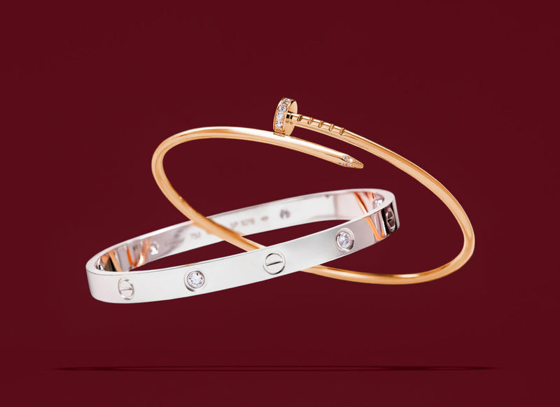 Two slim bangle bracelets overlapping on a deep burgundy background: one polished silver-toned bangle with screw details and clear stones, and one rose-gold-toned bangle with a screw detail and a small clear stone at the clasp.