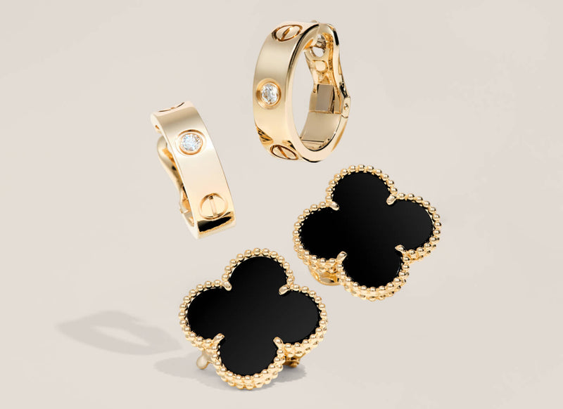 A luxury jewelry set featuring gold-plated huggie hoop earrings, stud earrings, and clover-shaped earrings. The earrings have black enamel centers with gold beaded borders, and the hoops and earrings feature small round clear gemstones.