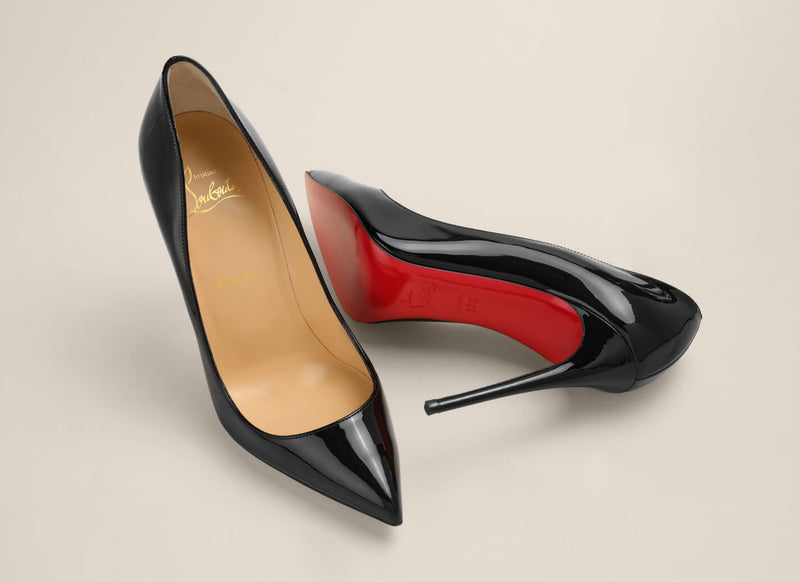 A pair of Christian Louboutin black patent leather pointed-toe pumps, showing the glossy black upper and the signature red outsole, set against a warm beige background.