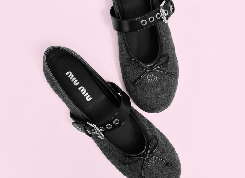 A pair of Miu Miu Mary Jane shoes in dark gray felt with black straps, shown from a top-down view on a solid light pink background.