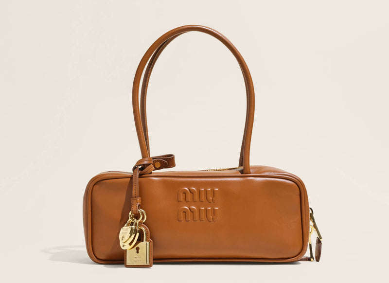 A Miu Miu brown leather shoulder bag with a top handle, gold-tone padlock charm, and embossed logo on the front, set against a light beige background.