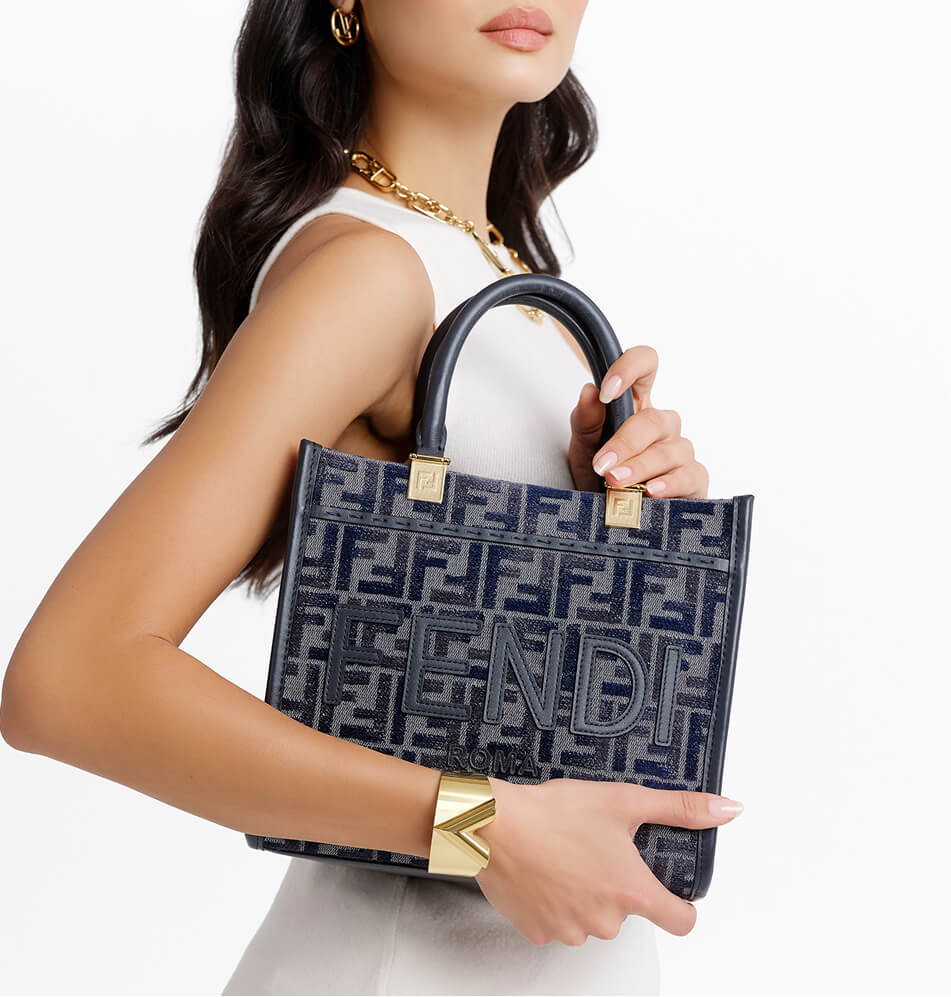 A woman holds a Fendi handbag with a navy blue and gray monogram pattern and gold-tone hardware against a clean white background.