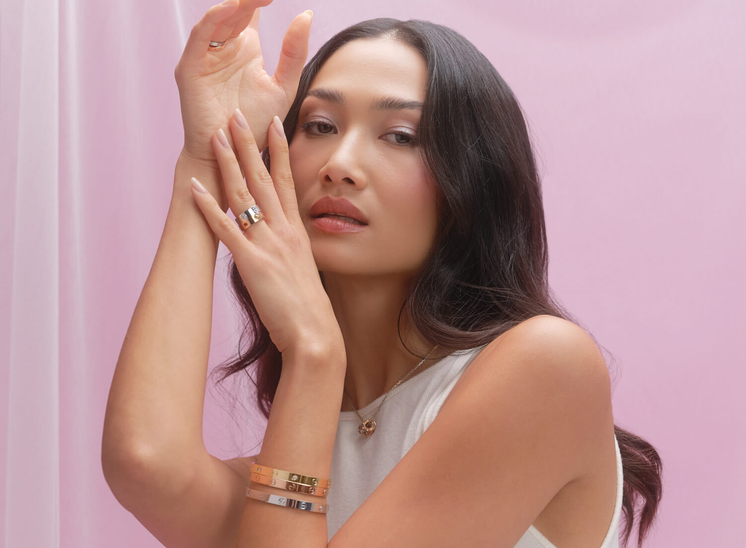 a woman wearing a white sleeveless top and holding her arms up showing three Cartier Love bracelets on her arm and two Cartier Love rings on her hand