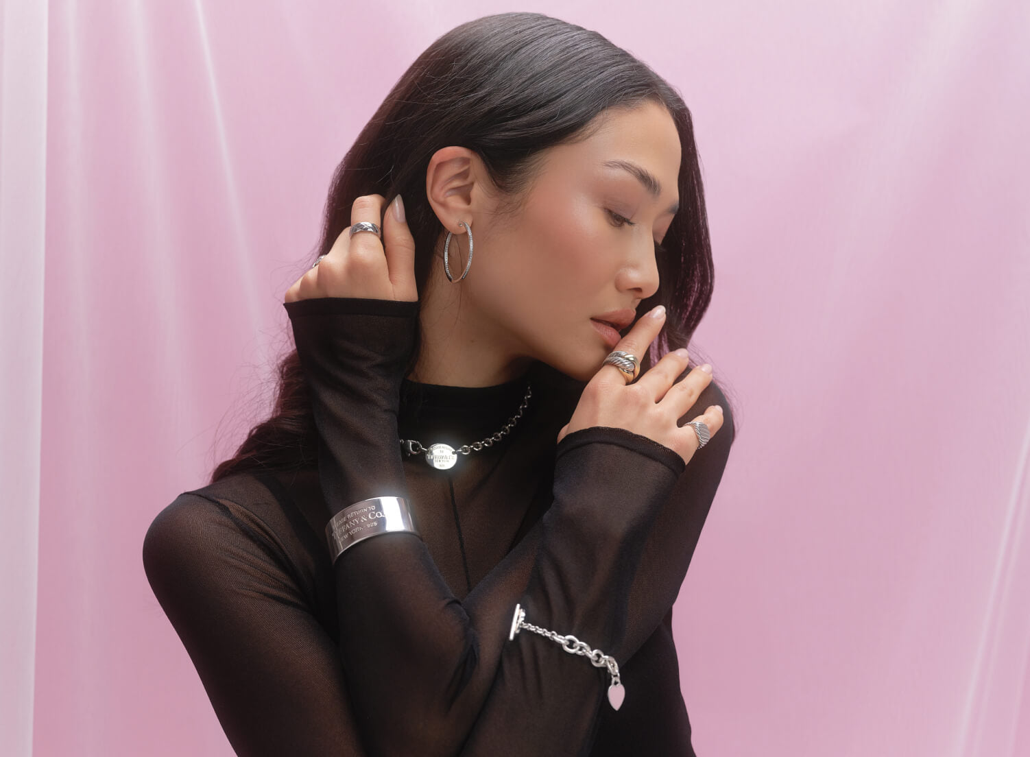 a woman wearing a black sheer mock neck long sleeve dress, a silver Return to Tiffany cuff bracelet, a silver Return to Tiffany heart charm bracelet, and a silver Return to Tiffany oval pendant charm necklace