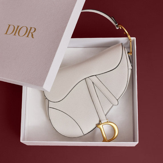 a white Christian Dior saddle bag with gold tone hardware laying in it's white box with Dior in gold on the lid sitting on a burgundy background