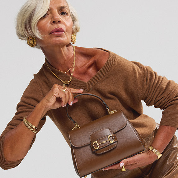 Grece Ghanem wearing a tan v-neck sweater and a brown leather midi skirt holding a brown Gucci Glazed Textured Calfskin Mini Horsebit 1955 Top Handle Flap Bag