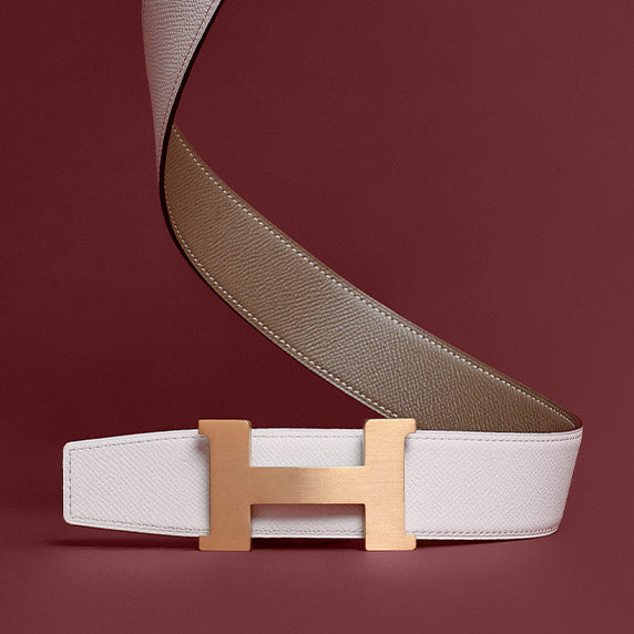 an Hermes white Constance Belt with a rose gold H shaped buckle sitting in front of a burgundy background
