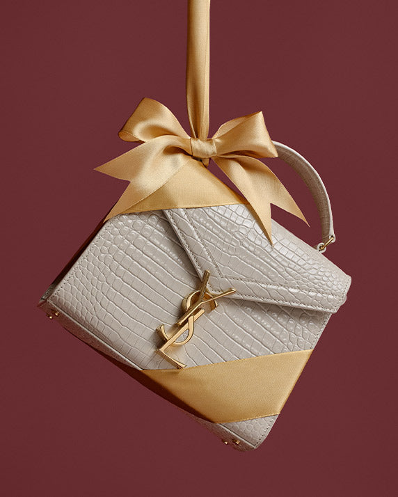 a cream color Saint Laurent Crocodile Embossed Mini Classic Monogram Cassandra Top Handle bag hanging by gold satin ribbon in front of a burgundy background