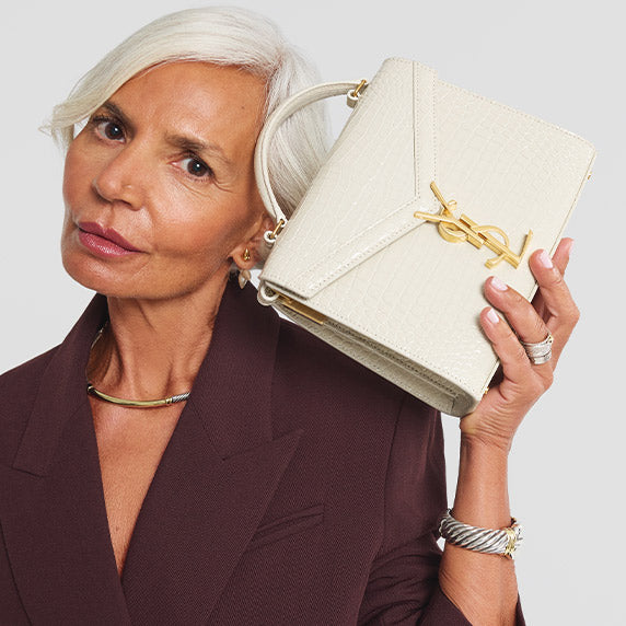 Grece Ghanem wearing a burgundy suit and holding a Saint Laurent cream color Crocodile Embossed Mini Classic Monogram Cassandra Top Handle bag next to her face
