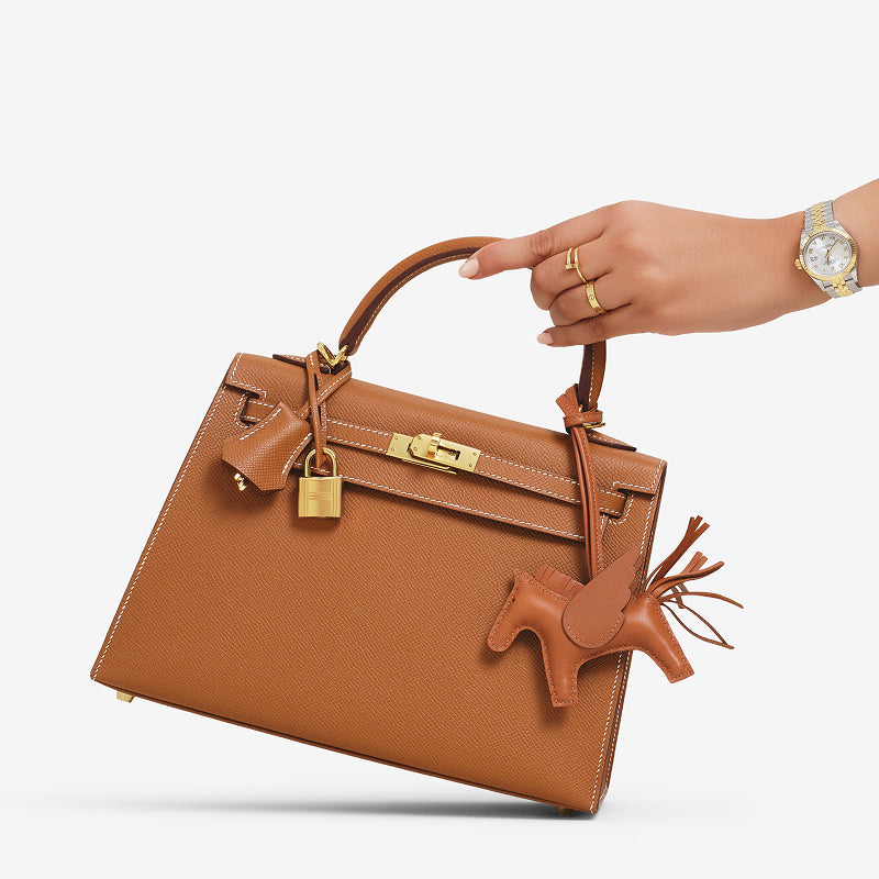 a woman's hand holding a Hermes Kelly 25 in a tan color with a Hermes Rodeo bag charm in a tan color hanging from the handle