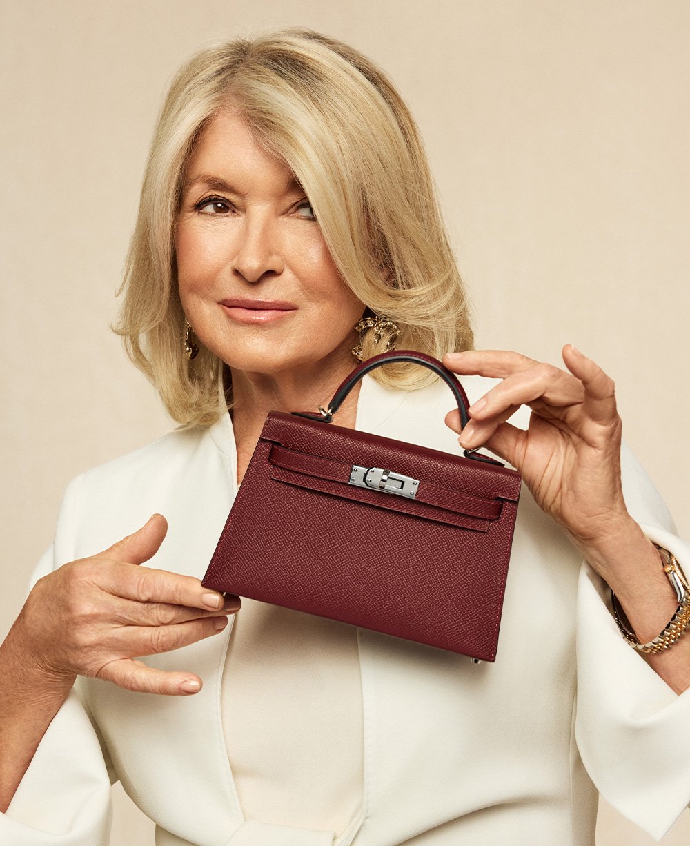 Martha Stewart wearing a while blazer and shirt and holding a burgundy color Hermes mini Kelly bag