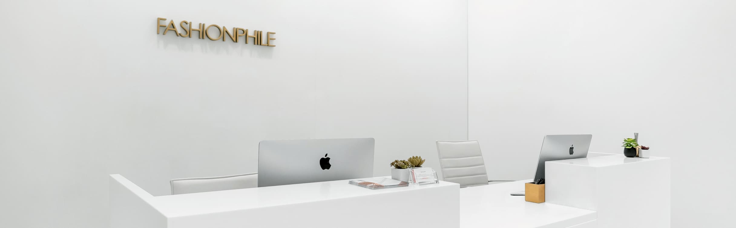 The modern, white reception area of a FASHIONPHILE Selling Studio, with the gold brand logo on the wall.