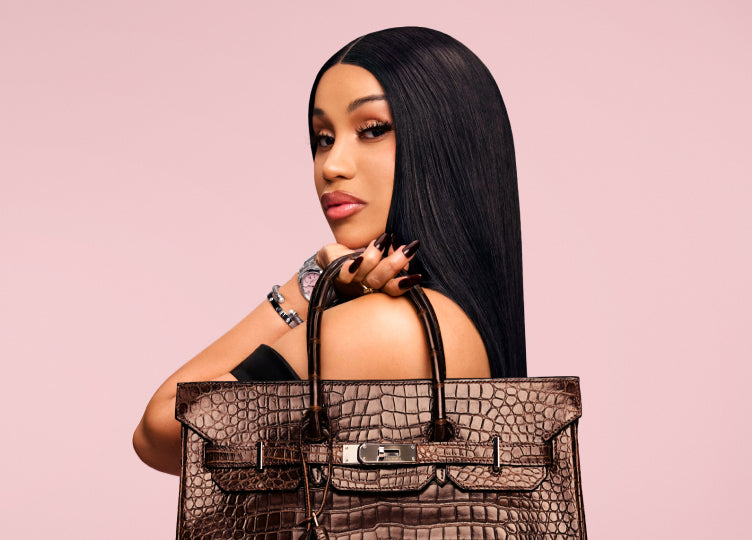A woman with long black hair holds a brown woven handbag against a solid dusty pink background.