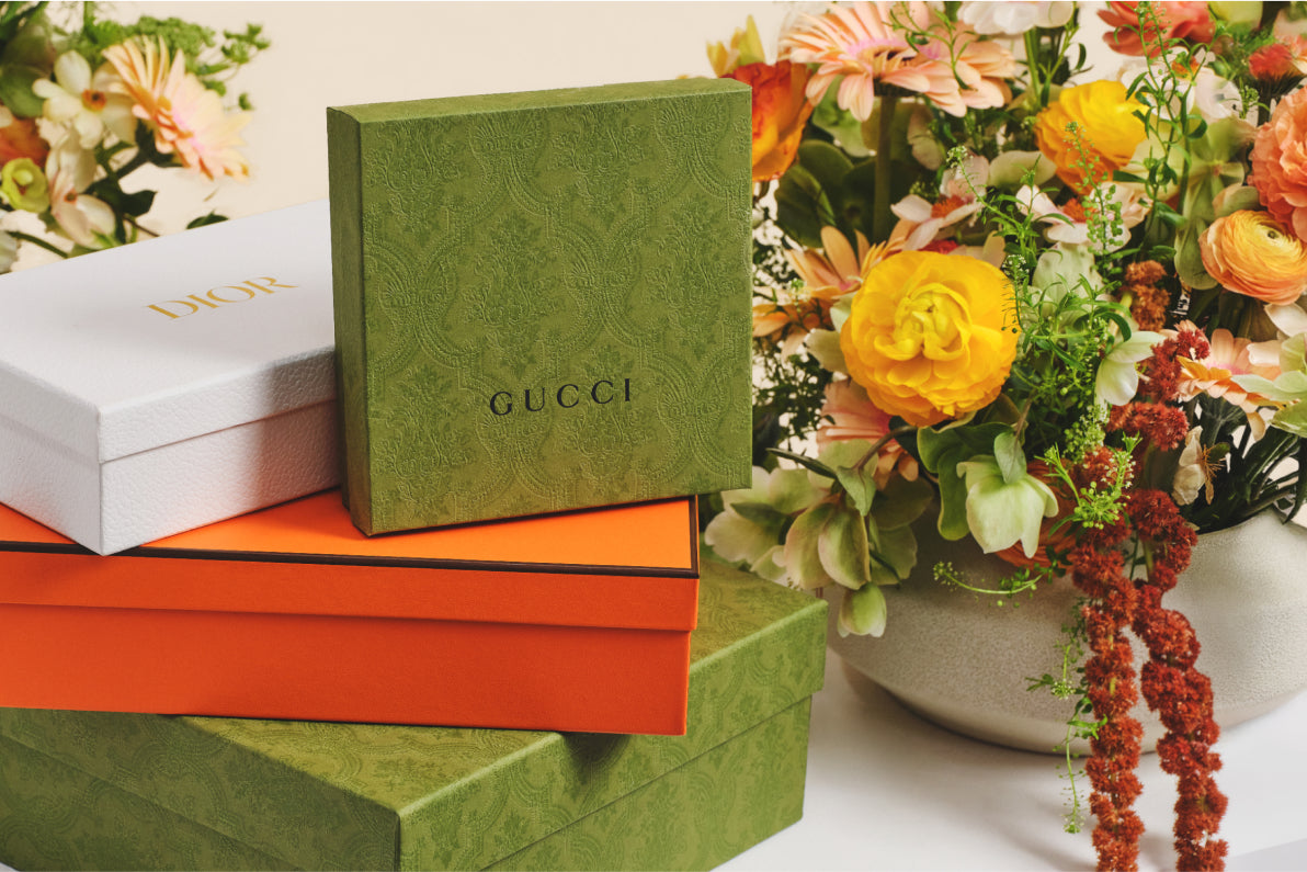 one green Gucci designer box and one white Christian Dior designer box sitting on top of one orange Hermes designer box on top of one green Gucci designer box with flowers in the background