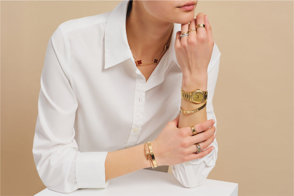 a woman in a white button down long sleeve shirt wearing a VAN CLEEF & ARPELS Gold Carnelian 10 Motifs Vintage Alhambra Necklace, a gold Rolex, a Cartier gold Juste Un Clou Bracelet, a Cartier Love Bracelet, and a TIFFANY Gold Atlas Bangle Bracelet