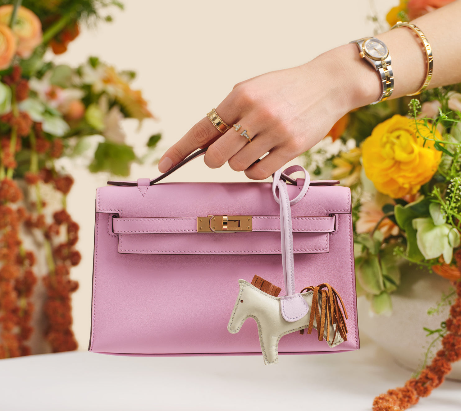 a woman's arm holding a HERMES Swift Kelly Pochette Clutch Mauve Sylvestre with a white Hermes Rodeo charm hanging on it with flowers in the background