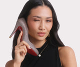 a woman wearing a black sleeveless dress and holding a glitter Christian Louboutin Kate heel against her face pretending it's a phone