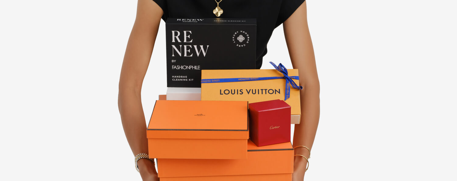 a woman wearing a black short sleeve dress and holding two orange Hermes boxes, one red Cartier box, and one orange Louis Vuitton box stacked together