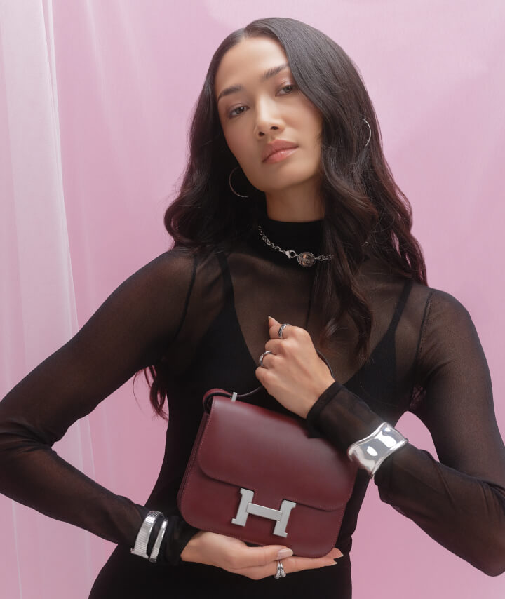a woman wearing a black sheer mock neck dress, a silver Tiffany bone cuff braacelet, and holding a Hermes Constance Rouge H color bag
